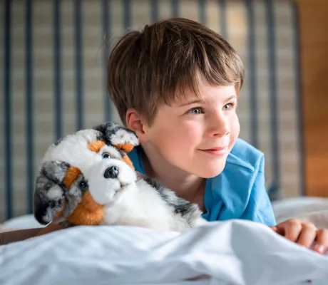 Ein Junge liegt in einem Bett im Familienhotel Sonnenalp Resort und hält ein Hunde-Stofftier
