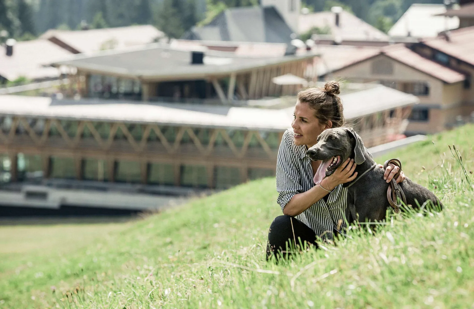 Eine Frau streichelt einen Hund auf einer Wiese vor dem Hundehotel HUBERTUS Mountain Refugio Allgäu sitzend
