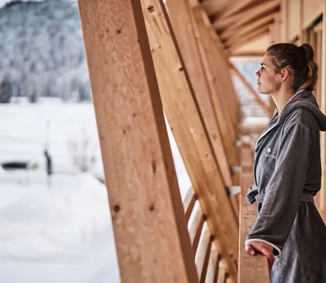 Eine Frau im Bademantel lehnt sich über ein Holzgeländer im Wellnesshotel HUBERTUS Mountain Refugio Allgäu