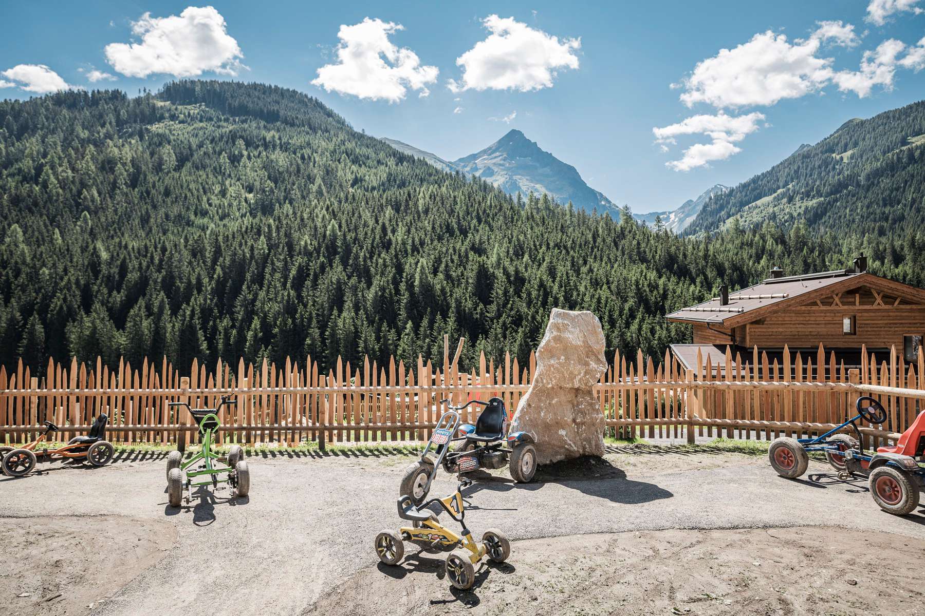 umzäunter Spielplatz mit Kinderfahrgeräten und Blick auf die Berge_FarmResortGeislerhof
