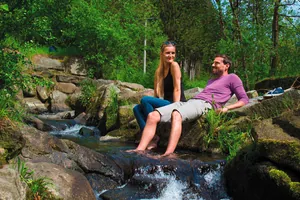 Eine Frau und ein Mann sitzen am Bach und der Mann hat seine Füße im Wasser