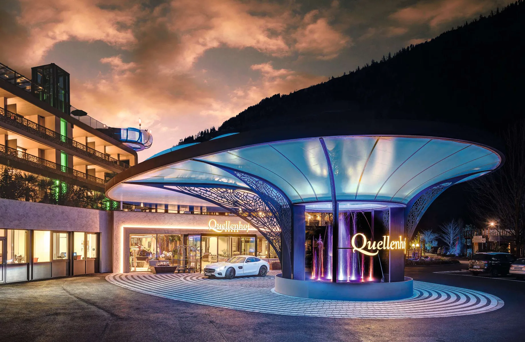 imposanter Eingangsbereich mit Wolkenspiel und weißem Auto vor dem Quellenhof Passeier