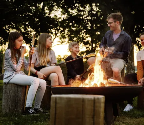 Mehrere Kinder sitzen zusammen mit einem Erwachsenen um eine Feuerschale und backen Stockbrot