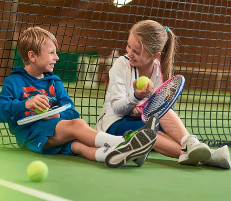 Zwei Kinder in Sportoutfits sitzen mit Tennisschlägern und -bällen vor Tennisnetz