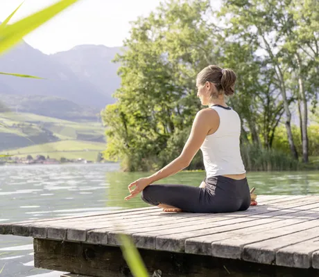 Frau sitzt auf Steg am See und macht Yoga