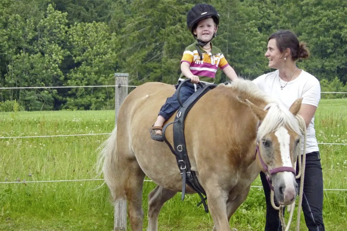 Betreuerin f&uuml;hrt Pferd mit kleinem Jungen mit Reithelm im Baumhaushotel Allg&auml;u