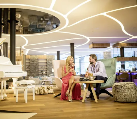 Frau in langem roten Kleid sitzt mit mann in der Lounge neben weißem Flügel in der Andreus Golf Lodge