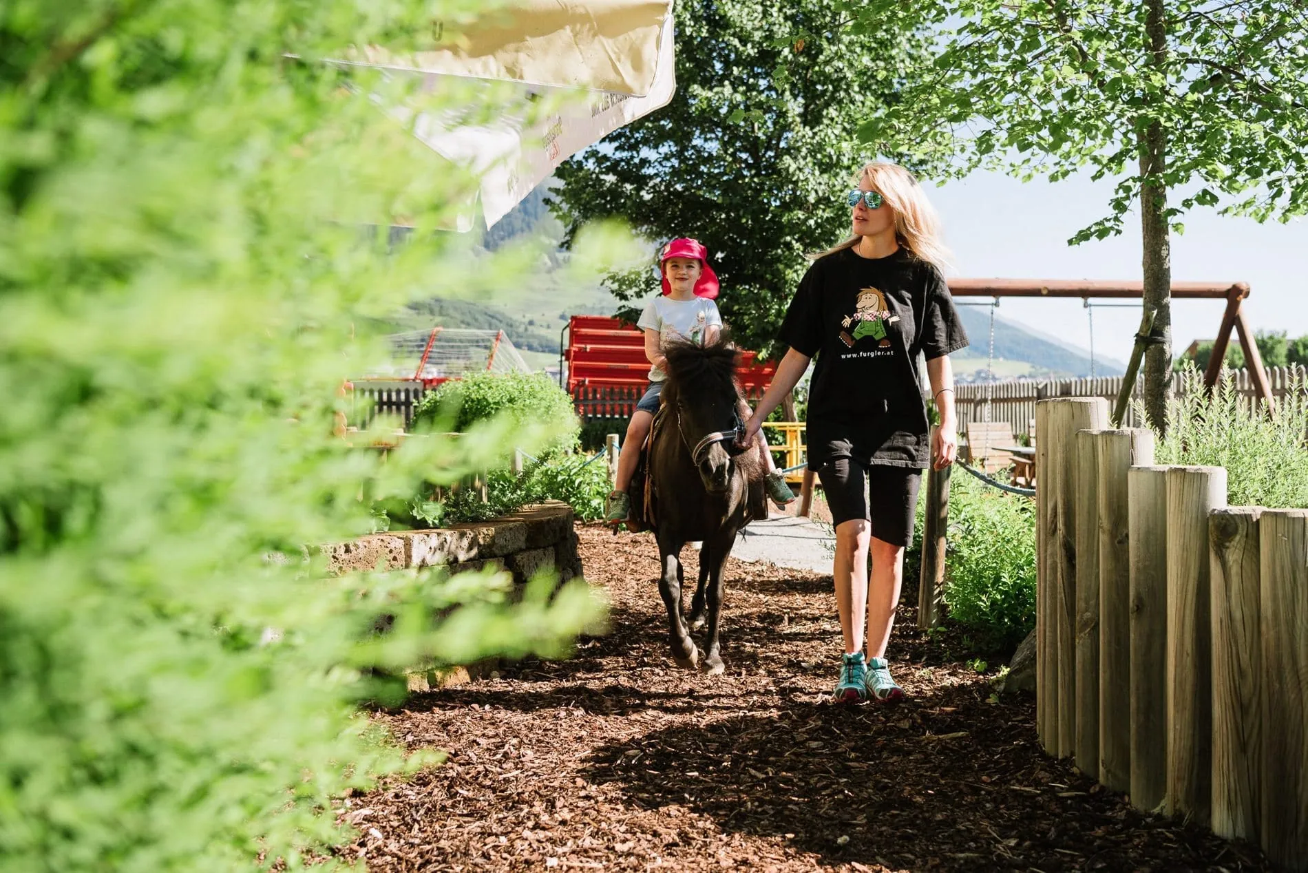 Junge Frau f&uuml;hrt kleines M&auml;dchen beim Ponyreiten durch die Hotelanlage