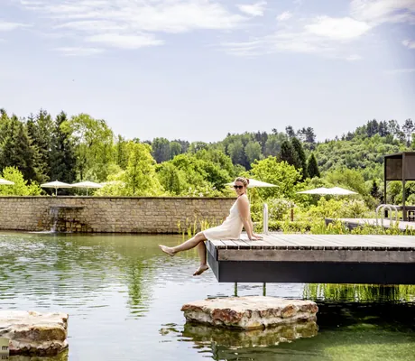 Frau im weißen Sommerkleid sitzt barfuß auf dem Steg am Badeteich im Pfalzblick Wald Spa Resort