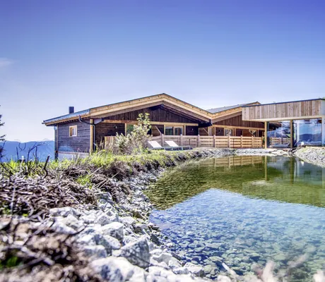 Blick auf das Chalet Stern und den benachbarten Wellnessbereich im Sommer mit Naturbadeteich im Vordergrund