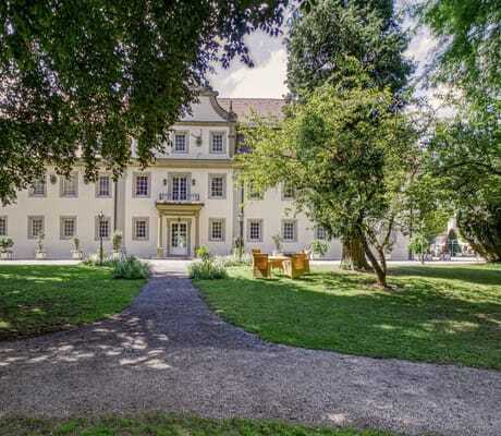 Blick vom Schlosspark auf die Schlossfassade des Wald- & Schlosshotel Friedrichsruhe mit Korbsitzgruppe davor