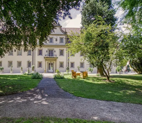 Blick vom Schlosspark auf die Schlossfassade des Wald- & Schlosshotel Friedrichsruhe mit Korbsitzgruppe davor