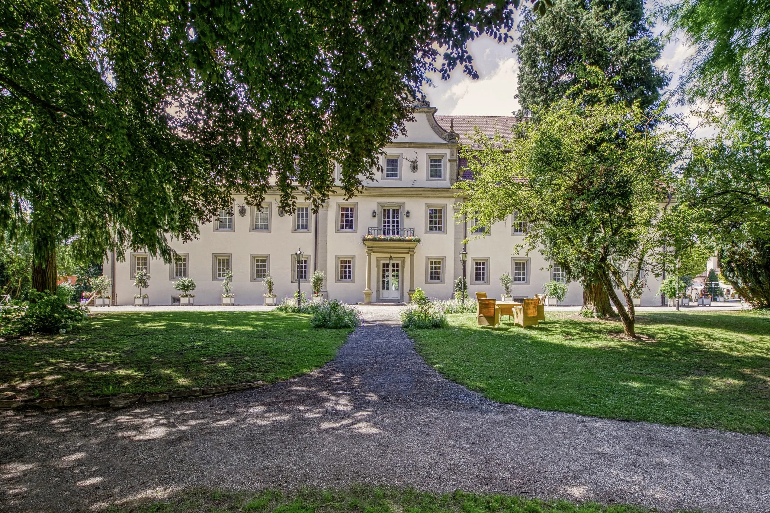 Blick vom Schlosspark auf die Schlossfassade des Wald- & Schlosshotel Friedrichsruhe mit Korbsitzgruppe davor