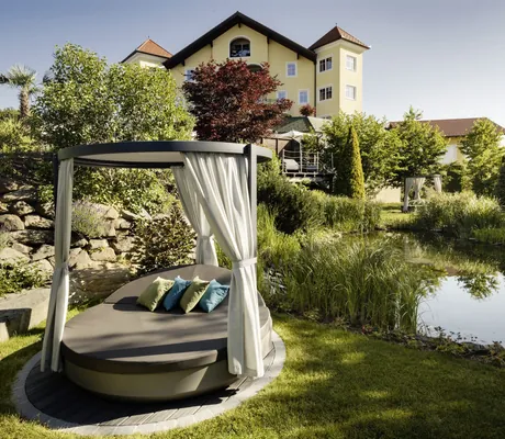 Himmelbett am Naturbadeteich vor gelbem Gebäude mit Türmchen im Wellnesshotel Jagdhof Röhrnbach
