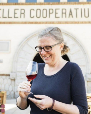 Autorenporträt Heidi Siefert in blauem Shirt mi Brille ein Weinglas haltend vor einem Schild 'Destiller Cooperatiu'