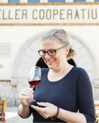 Autorenporträt Heidi Siefert in blauem Shirt mi Brille ein Weinglas haltend vor einem Schild 'Destiller Cooperatiu'