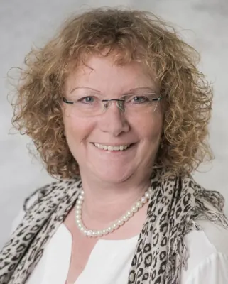 Autorenporträt Karin Gabler mit Brille lächelnd mit weißer Bluse, Perlenkette und Tiger-Print-Schultertuch