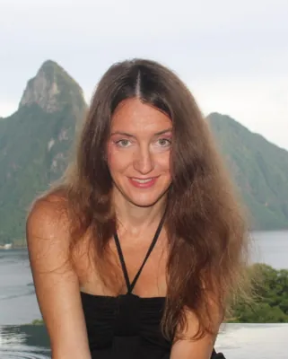 Autorenporträt Manuela Blisse sitzt lächelnd mit offenem Haar in schwarzem Top vor See und Bergkulisse