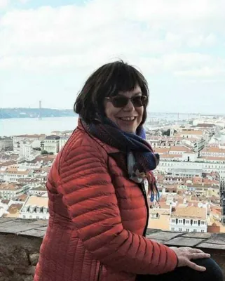 Autorenporträt Martina Schäfer steht lächelnd mit Sonnenbrille und Anorak vor Stadtkulisse