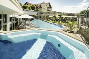 Blick vom Whirlpool auf den Außenpool und gelbes Gebäude mit Türmchen im Wellnesshotel Jagdhof Röhrnbach