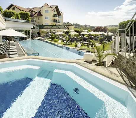 Blick vom Whirlpool auf den Außenpool und gelbes Gebäude mit Türmchen im Wellnesshotel Jagdhof Röhrnbach