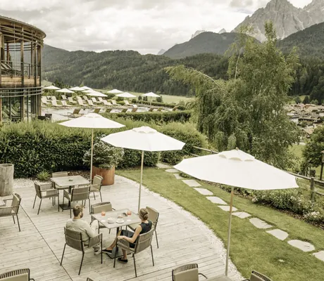 Paar unter beigem Sonnenschirm auf der Holzterrasse des Naturhotel Leitlhof genießt den Ausblick auf die Dolomiten