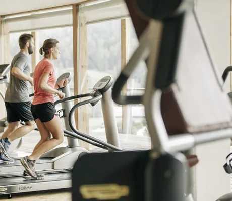 Frau in rotem T-Shirt und Mann in grauem T-Shirt laufen auf dem Laufband im Fitnessraum des Naturhotel Leitlhof