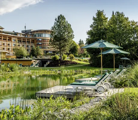 Naturbadeteich mit grün gepolsterten Liegen mit Blick auf die Holzfassade des Sonnenalp Resort