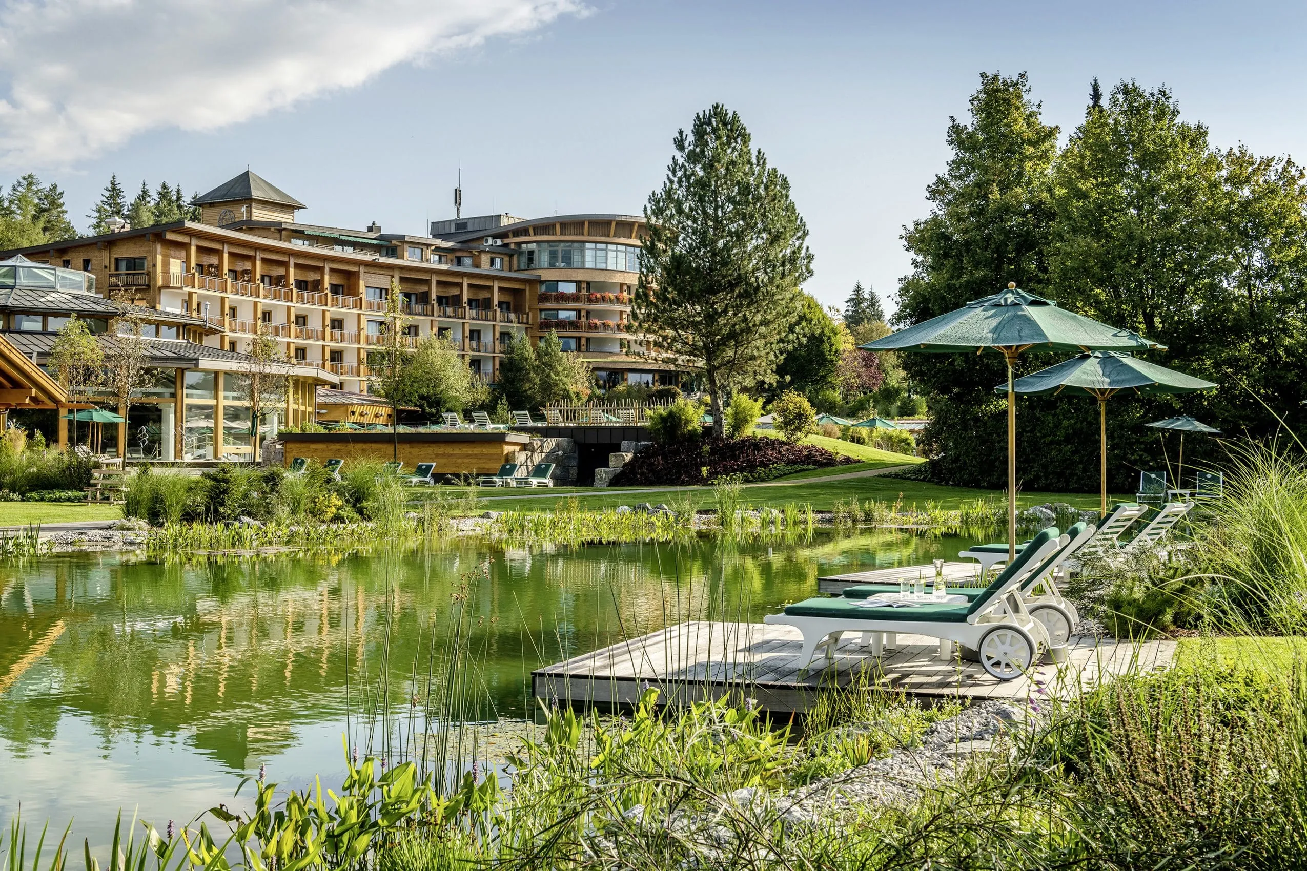 Naturbadeteich mit gr&uuml;n gepolsterten Liegen mit Blick auf die Holzfassade des Sonnenalp Resort