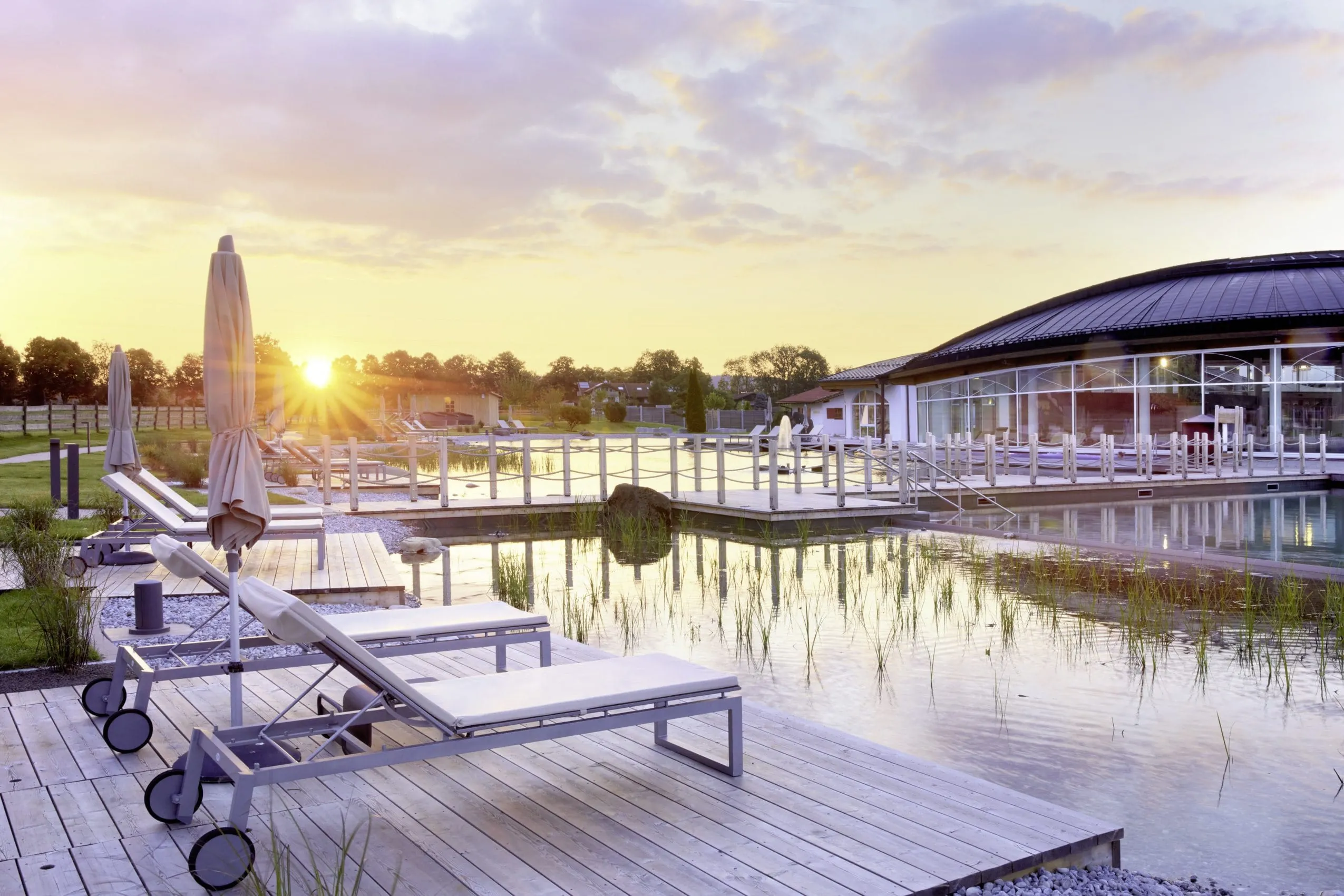 Liegen auf Holzpontons am Naturbadeteich mit Blick auf das ovale Wellnessgeb&auml;ude des K&ouml;nig Ludwig Inspiration Spa