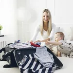 Mutter packt Koffer auf dem Bett, während Baby daneben krabbelt