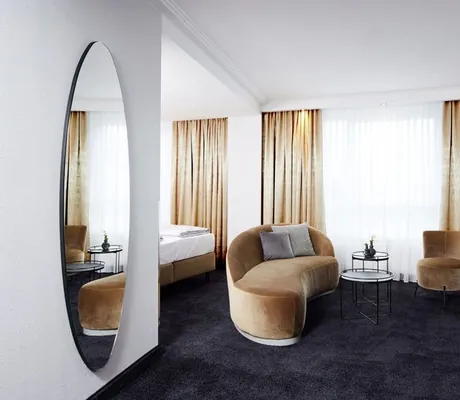 Junior-Suite in Loungeästhetik mit beiger Samt-Bestuhlung und -vorhängen auf schwarzem Teppichboden