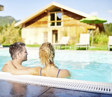 Rückenansicht eines Paares im Outdoorpool mit Blick auf das Lodge- und Tagungsgebäude im Hotel Oberstdorf