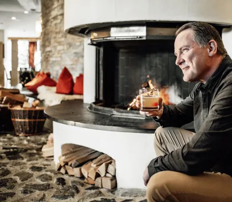 Männlicher Hotelgast sitzt lächelnd mit Drink am offenen Kamin der Pianobar