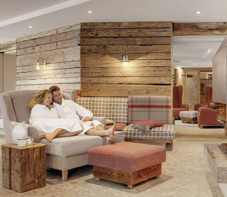 Paar in Bademänteln kuschelt in einem Oversize-Sessel im Wellness-Wohnzimmer am offenen Kaminfeuer des Hotel Oberstdorf