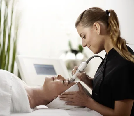 Kosmetikerin arbeitet mit Kosemtikgerät im Gesicht einer Frau mit Frotteeturban im Esperanto