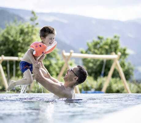 Vater mit Brille wirft kleinen Jungen mit Schwimmflügeln im Pool durch die Luft