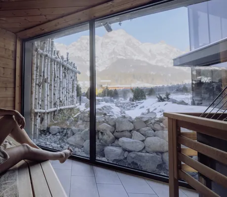 Junge Frau sitzt im Winter in der Sauna und blickt durch Panoramafenster in die Berge