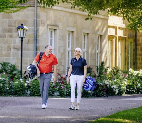Zwei Personen mit Golf-Equipment laufen Richtung Golfplatz des Gräflicher Park Health & Balance Resort