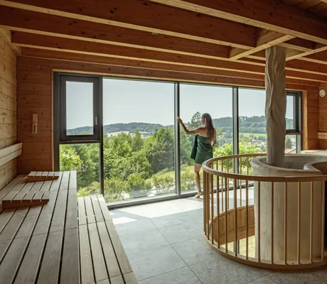 Frau in grünem Handtuch steht am Fenster der Sauna mit rundem Saunaofen in der Mitte