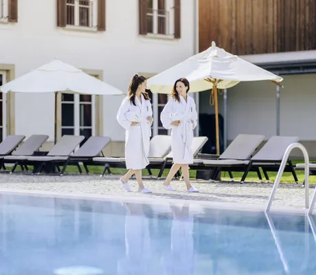 zwei drunkelhaarige Frauen in weißen Bademänteln laufen am Beckenrand des Outdoorpools entlang