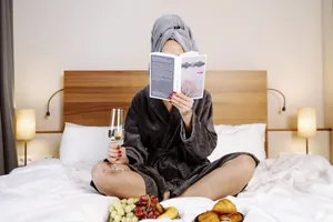 Frau in anthrazitfarbenem Bademantel mit Frotteeturban hockt im Schneidersitz auf dem Bett und liest