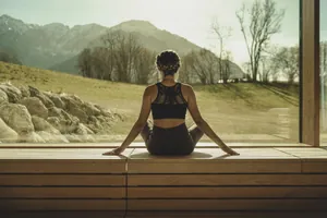 Frau in dunklem Gymnastikaoutfit und mit geflochtenem Haarkranz sitzt im Schneidersitz vor den Panoramafenster