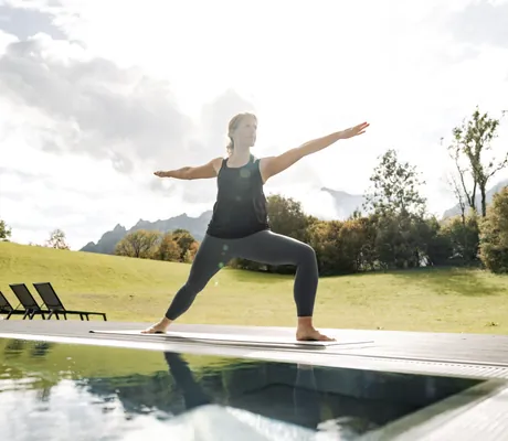 blonde Frau mit Zopf steht in Gymnastikoutfit in Kriegerhaltung am Outdoorpool des Klosterhof Bayerisch Gmain