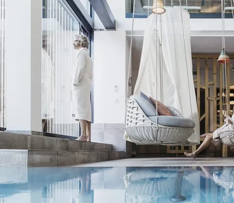 Mann im Bademantel steht am Panoramafenster, während Frau in Hängekorbsessel am Pool schaukelt