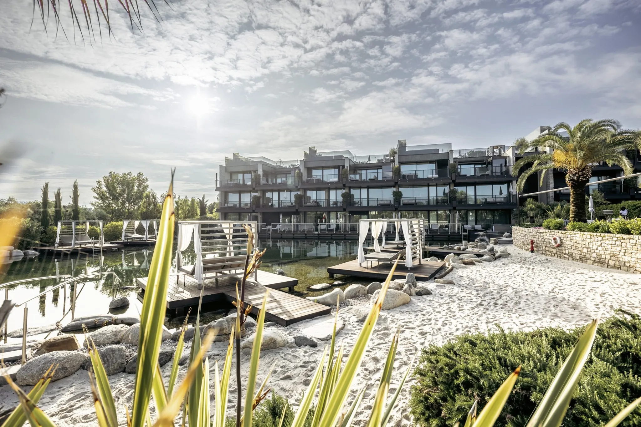 Strand mit Holzstegen auf den Naturbadeteich und Blick auf die Fassade des Quellenhof Luxury Resort Lazise