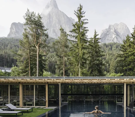 Frau mit langen Haaren steht mit ausgebreiteten Armen im Outdoorpool mit Blick auf den Schlern