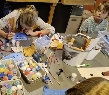 Kinder bemalen blaue selbstgebastelte Katzengesichter an einem Tisch in der Kinderwerkstatt Berghaus Schröcken