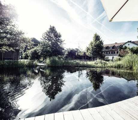 See mit geschwungener Holzterrasse Sonnenschirm und Blick auf Ökonomiegebäude im Schlossgut Oberambach in Münsing