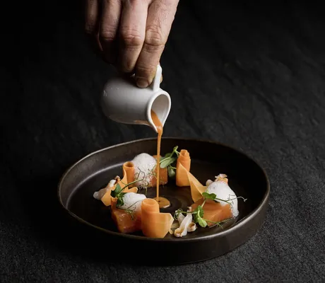 orange-rote Soße wird aus einem weißen Kännchen auf ein Lachsgericht gegossen im La Maiena Meran Resort